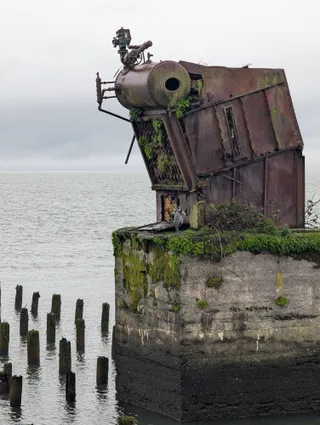 A rusting old boiler on a moss-covered concrete pier, with a great blue heron sheltering beneath it