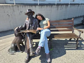 Euri leaning on the shoulder of a bronze statue of a bearded man reading a newspaper, with a bronze dog beside him